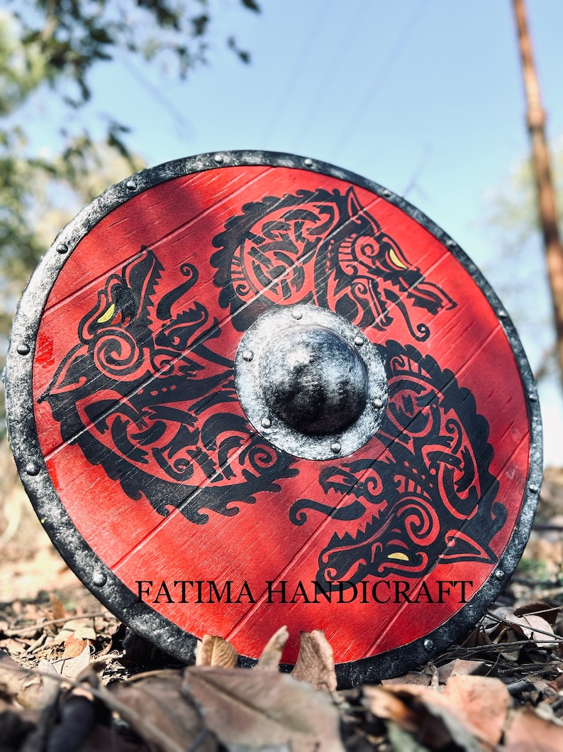 K&ouml;nnte beinhalten: Ein roter Holzschild mit einem schwarz-wei&szlig;en Design von zwei W&ouml;lfen. Der Schild hat einen Metallrand und einen Metallboss in der Mitte. Der Schild liegt auf einem Bett aus braunen Bl&auml;ttern. Der Text "FATIMA HANDICRAFT" ist auf der Unterseite des Schildes aufgedruckt.