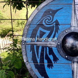 K&ouml;nnte beinhalten: Ein blauer Holzschild mit einer schwarzen Silhouette eines Vogels mit einem Spiralmuster. Der Schild hat einen Metallrand mit Nieten und einen Metallboss in der Mitte. Der Text "Fatima Handicraft" ist auf dem Schild sichtbar.