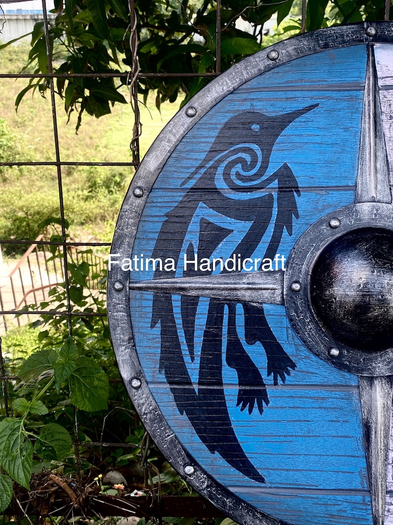 K&ouml;nnte beinhalten: Ein blauer Holzschild mit einer schwarzen Silhouette eines Vogels mit einem Spiralmuster. Der Schild hat einen Metallrand mit Nieten und einen Metallboss in der Mitte. Der Text "Fatima Handicraft" ist auf dem Schild sichtbar.
