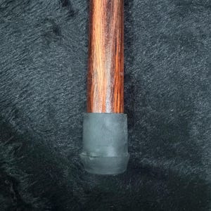 Op de afbeelding: Een houten wandelstok met een donkerbruin en roodbruin nerfpatroon. De onderkant van de stok heeft een zwarte rubberen dop voor stabiliteit. De stok is ontworpen voor ondersteuning en mobiliteit.