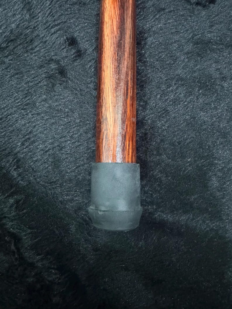 Op de afbeelding: Een houten wandelstok met een donkerbruin en roodbruin nerfpatroon. De onderkant van de stok heeft een zwarte rubberen dop voor stabiliteit. De stok is ontworpen voor ondersteuning en mobiliteit.
