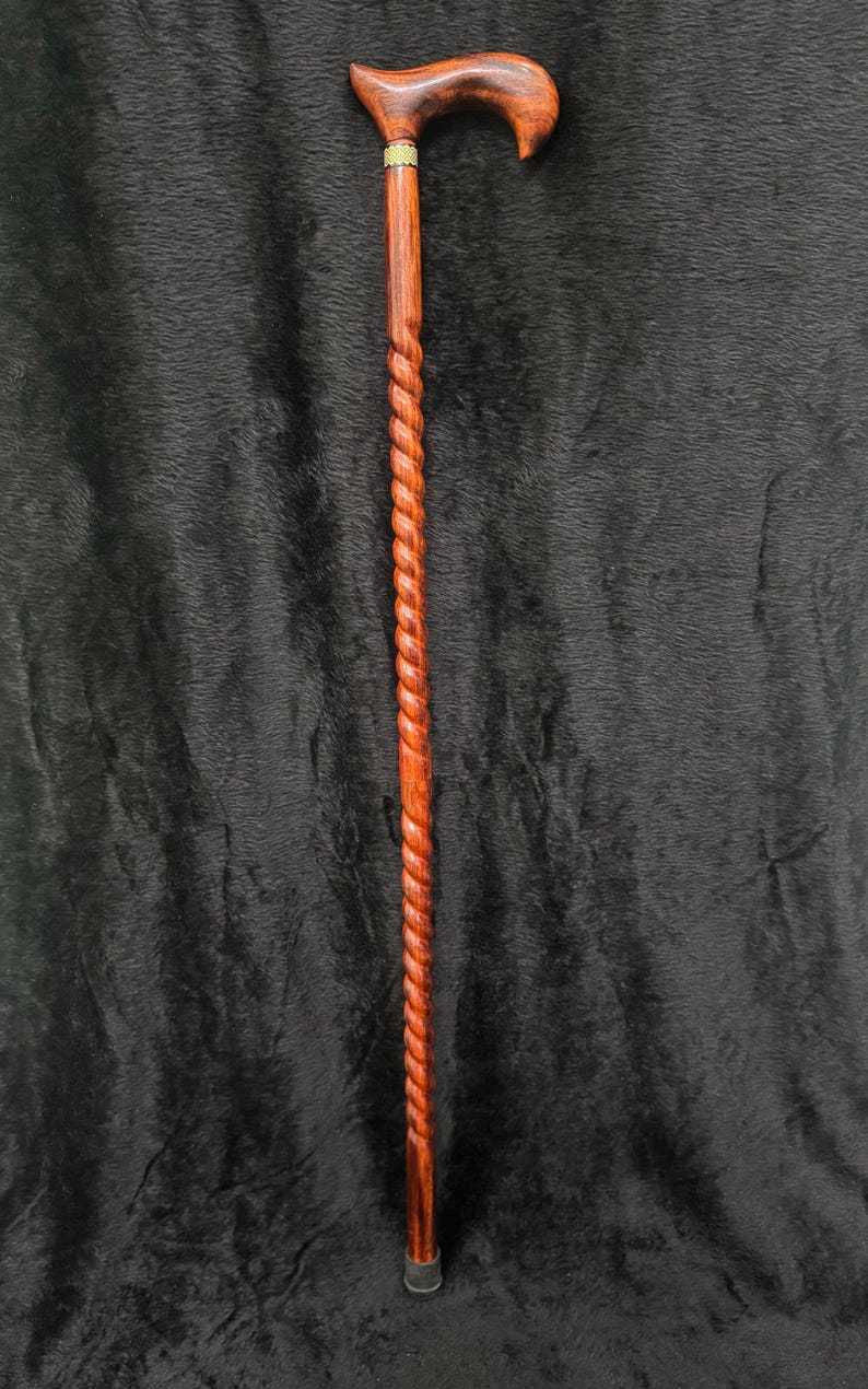 Op de afbeelding: Een houten wandelstok met een gebogen handvat en een gedraaide schacht. De stok is bruin van kleur met een messing accent bij het handvat. De stok heeft een rubberen dop.