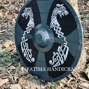K&ouml;nnte beinhalten: Ein schwarzer und silberner Holzschild mit zwei darauf gemalten Drachenk&ouml;pfen. Der Schild ist mit komplizierten Knotenarbeiten verziert und hat einen Metallrand. Der Schild liegt auf dem Boden mit Bl&auml;ttern und &Auml;sten drumherum. Der Text "FATIMA HANDICRAFT" ist am unteren Rand des Schildes sichtbar.