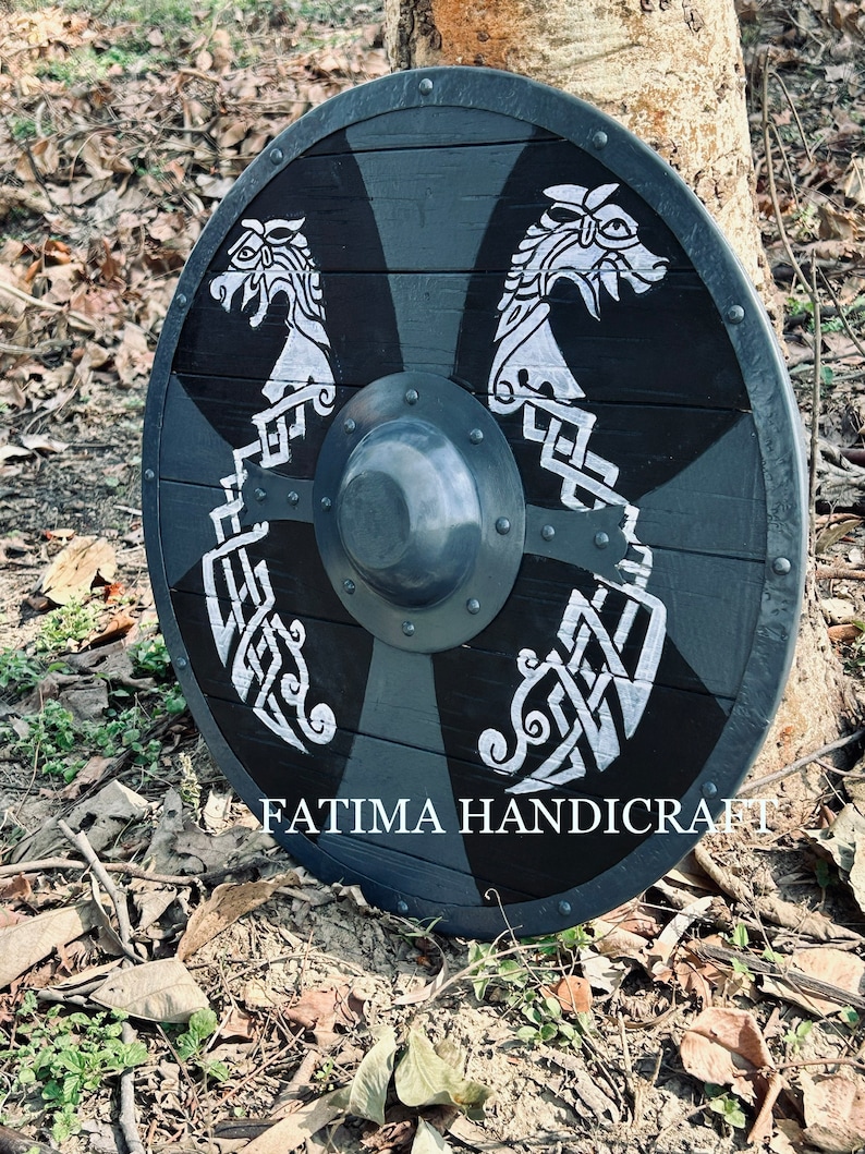 K&ouml;nnte beinhalten: Ein schwarzer und silberner Holzschild mit zwei darauf gemalten Drachenk&ouml;pfen. Der Schild ist mit komplizierten Knotenarbeiten verziert und hat einen Metallrand. Der Schild liegt auf dem Boden mit Bl&auml;ttern und &Auml;sten drumherum. Der Text "FATIMA HANDICRAFT" ist am unteren Rand des Schildes sichtbar.