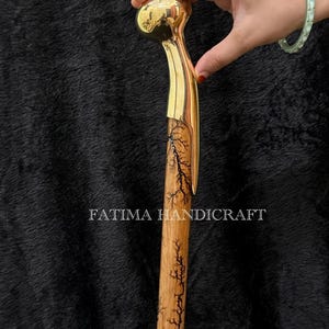 May include: A handcrafted walking cane with a polished brass handle and a wooden shaft. The shaft features a detailed tree branch design. The text "FATIMA HANDICRAFT" is printed on the shaft.