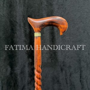 Op de afbeelding: Handgemaakte houten wandelstok met een gebogen handvat en een gedraaide schacht. De stok heeft een rijke bruine kleur met een goudkleurige band bij het handvat. De tekst "FATIMA HANDICRAFT" is op de stok gedrukt.