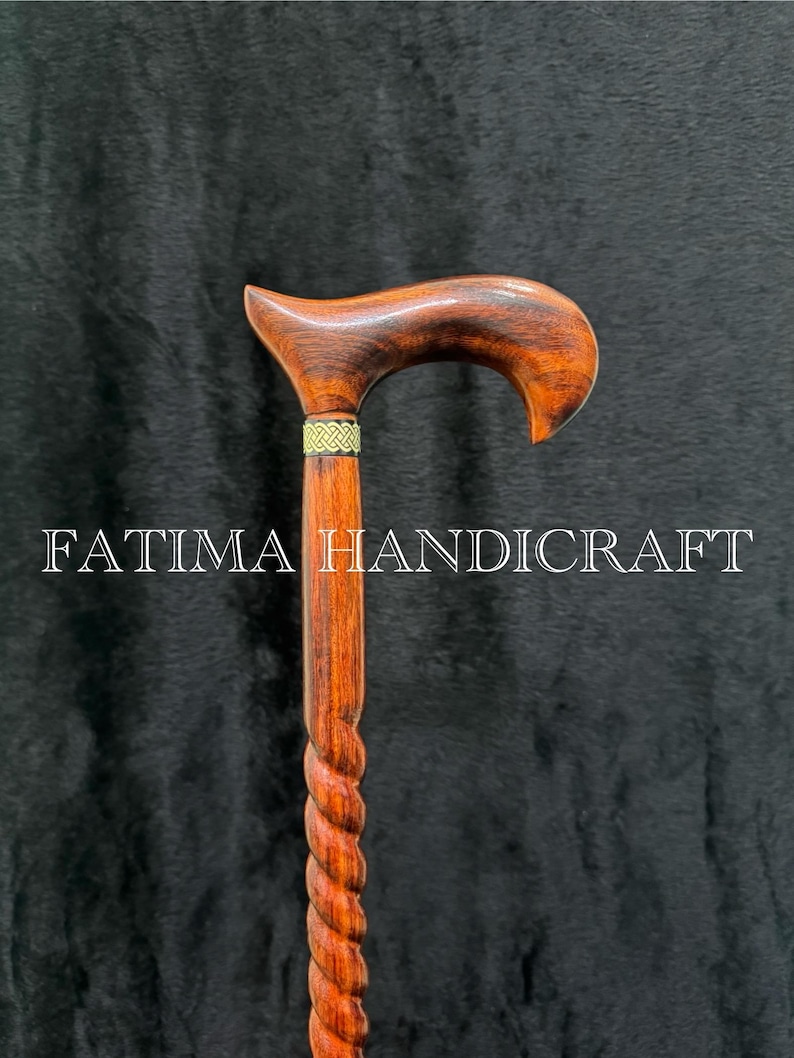 Op de afbeelding: Handgemaakte houten wandelstok met een gebogen handvat en een gedraaide schacht. De stok heeft een rijke bruine kleur met een goudkleurige band bij het handvat. De tekst "FATIMA HANDICRAFT" is op de stok gedrukt.