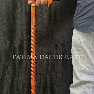 Op de afbeelding: Handgemaakte houten wandelstok met een gedraaid touwontwerp. De handgreep is van gebogen, gepolijst hout met een gouden accent. De tekst "FATIMA HANDICRAFT" is op de stok gedrukt.