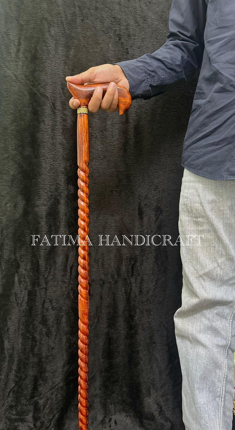 Op de afbeelding: Handgemaakte houten wandelstok met een gedraaid touwontwerp. De handgreep is van gebogen, gepolijst hout met een gouden accent. De tekst "FATIMA HANDICRAFT" is op de stok gedrukt.