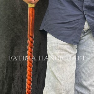 Op de afbeelding: Een houten wandelstok met een gedraaid ontwerp. De stok is diep roodbruin van kleur, met een goudkleurige band aan de bovenkant. De tekst "FATIMA HANDICRAFT" is zichtbaar op de afbeelding.