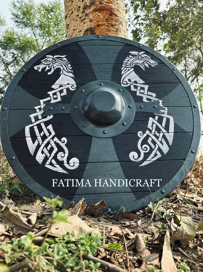 K&ouml;nnte beinhalten: Ein schwarz-wei&szlig;er Holzschild mit einem Metallrand und einem zentralen Boss. Der Schild ist mit zwei wei&szlig;en Drachenk&ouml;pfen und komplizierten Knotenmustern verziert. Der Text "FATIMA HANDICRAFT" ist auf dem Schild aufgedruckt.