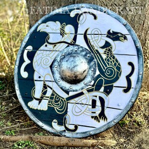 K&ouml;nnte beinhalten: Ein runder Holzschild mit einem schwarz-wei&szlig;en Design. Der Schild zeigt einen wei&szlig;en Drachen und einen schwarzen Vogel mit goldenen Akzenten. Der Schild ist mit Metallnieten am Rand verziert. Der Text "FATIMA HANDICRAFT" ist oben auf dem Schild sichtbar.