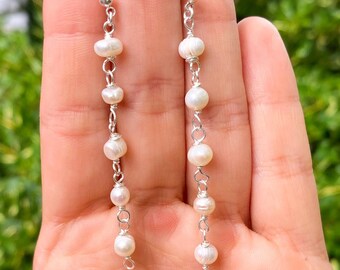 Puede incluir: Un par de pendientes colgantes con cinco pequeñas perlas de agua dulce blancas de forma irregular cada uno, unidas por un fino alambre de metal plateado y rematadas con un pequeño perno de bola plateado en la parte superior.
