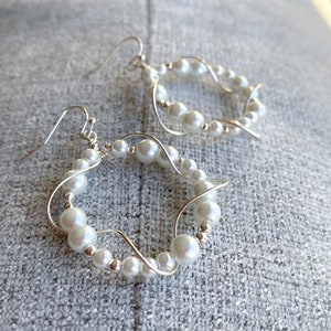 Può includere: Un paio di orecchini a cerchio d'argento con perle bianche. Gli orecchini sono realizzati in filo metallico e hanno un design delicato e femminile.