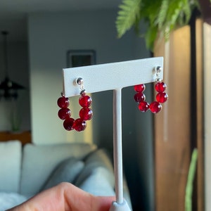 May include: A pair of red gemstone hoop earrings with silver posts. The earrings are displayed on a white stand.
