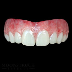 Può includere: Un set di denti superiori con smalto bianco e lucido e gengive rosa. I denti sono disposti in un arco naturale, con un piccolo spazio tra i due denti anteriori. Lo sfondo è nero. Il testo "MOONSTRUCK EFFECTS" è in basso.