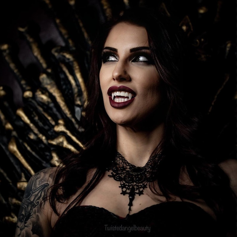 May include: A woman with dark hair and dark eye makeup is wearing a black lace choker and a black dress. She has fangs and is looking up with a serious expression. The background is a dark, textured surface.