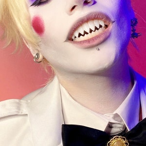 K&ouml;nnte beinhalten: Eine Person mit blonden Haaren und wei&szlig;em Make-up tr&auml;gt ein wei&szlig;es Hemd mit einer schwarzen Schleife und einer goldenen Brosche. Sie hat einen Nasenring und ein Lippenpiercing. Ihre Z&auml;hne sind spitz und zugespitzt.