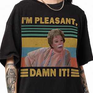 May include: A black t-shirt with a vintage-style graphic featuring a woman with a surprised expression. The graphic includes the text "I'm Pleasant, Damn It!" and a retro-style striped background.