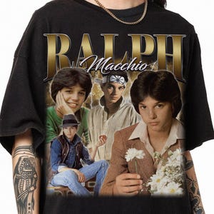 May include: Black t-shirt featuring a collage of images of Ralph Macchio with the name "RALPH Macchio" in gold lettering. The design includes multiple images of the actor in various poses, with a vintage aesthetic.
