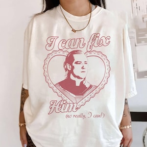 May include: Cream-colored t-shirt with a pink graphic. The graphic features a portrait inside a heart-shaped frame, with the text "I can fix Him" and "(no really, I can!)". The shirt has short sleeves and a crew neck.