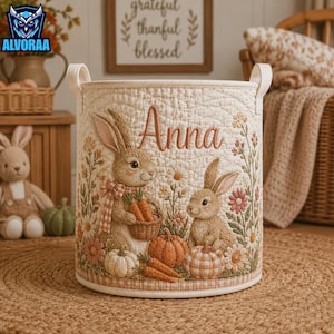 May include: A cylindrical fabric storage basket with a quilted cream exterior featuring an embroidered design of two brown rabbits, pumpkins, and flowers. The name Anna is stitched in brown script above the rabbits, and the basket has two handles.