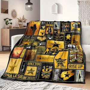 May include: A black and gold blanket featuring various graphics and text from the musical "Hamilton." The blanket displays phrases like "Rise Up," "Talk Less," and "I Am Not Throwing Away My Shot." The design includes star and silhouette imagery.