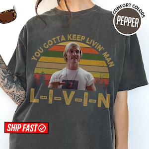 May include: Dark gray t-shirt with a graphic featuring a retro sunset design and the phrase "You Gotta Keep Livin' Man." The image includes a portrait of a man and the word "L-I-V-I-N" in yellow block letters. The shirt has a crew neck.