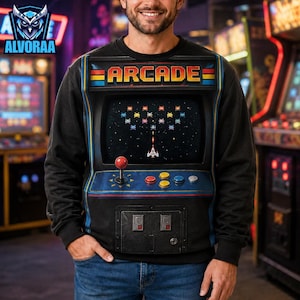May include: A black long-sleeve sweatshirt featuring a large graphic print of a vintage arcade cabinet. The design displays the word ARCADE in yellow letters, a screen showing a space shooter game with colorful pixelated enemies, a red joystick, and buttons.