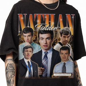 May include: Black t-shirt featuring a graphic of Nathan Fielder with the text "NATHAN Fielder" in a retro style. The design includes multiple images of Nathan Fielder. The shirt has a vintage aesthetic.