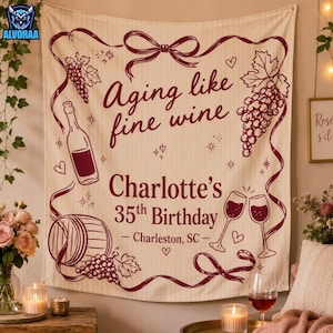 May include: A cream-colored fabric wall hanging featuring a maroon graphic design of a wine bottle, a barrel, grape clusters, and two wine glasses. The text reads: Aging like fine wine, Charlotte's 35th Birthday, Charleston, SC, with ribbons.