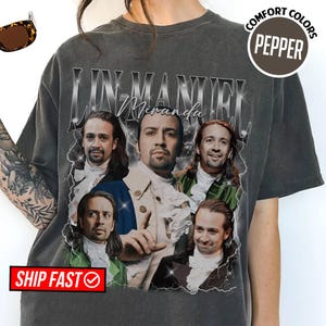 May include: Dark gray t-shirt featuring a graphic of Lin-Manuel Miranda in various poses. The design includes the name "Lin-Manuel Miranda" in a stylized font, with a vintage aesthetic. The shirt has a crew neck and short sleeves.