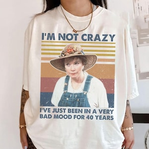 May include: Cream-colored t-shirt with a graphic featuring a woman in a straw hat and denim overalls. The text reads "I'M NOT CRAZY" above the image and "I'VE JUST BEEN IN A VERY BAD MOOD FOR 40 YEARS" below.