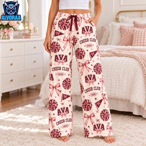 May include: Cream-colored pajama pants featuring a repeating pattern of maroon cheerleading pom-poms, bows, pennants, and the text "AVA CHEER CLUB 2026". The pants have a wide-leg silhouette and a maroon drawstring waist tie.