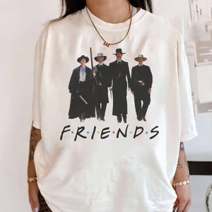 May include: White t-shirt with a graphic of five men in cowboy hats and the text "FRIENDS" in colorful letters.