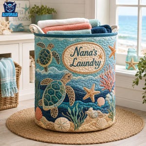May include: A cylindrical fabric laundry hamper featuring a quilted ocean theme with sea turtles, starfish, coral, and seashells. The front displays an oval patch with the text Nana's Laundry. The hamper is filled with folded towels.