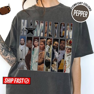 May include: Dark gray t-shirt featuring the word "HAMILTON" in a bold, silver font. The shirt showcases a star graphic and images of characters from the musical. The shirt also has the words "COMFORT COLORS" and "PEPPER" printed on it.
