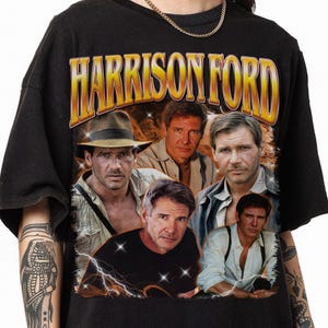 May include: Black t-shirt featuring a collage of images of Harrison Ford, with the name "HARRISON FORD" in a gold and orange font across the top. The images include Ford in various roles, with lightning bolt graphics.