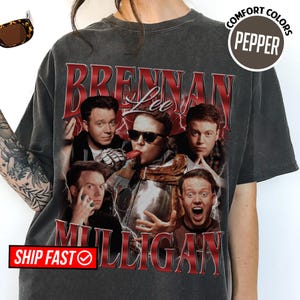 May include: Dark gray t-shirt featuring a collage of images and the text "Brennan Lee Mulligan" in red. The shirt has a vintage look with the words "Comfort Colors" and "Pepper" printed on it.