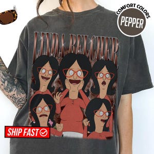 May include: Dark gray t-shirt featuring a graphic of Linda Belcher from Bob's Burgers. The design includes multiple images of Linda wearing red glasses and a red shirt. The text "LINDA BELCHER" is above the graphic. The shirt has a "COMFORT COLORS" label.