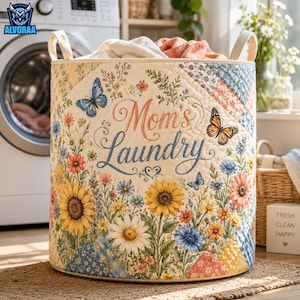 May include: A cylindrical quilted laundry basket featuring a cream-colored background with a vibrant floral print of sunflowers, daisies, and butterflies. The text "Mom's Laundry" is printed in the center in a decorative script font.