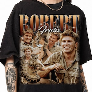 May include: Black t-shirt with a vintage-style graphic of Robert Irwin. The graphic features a collage of photos of Robert Irwin with animals, including a koala and a snake. The text "Robert Irwin" is printed in large, bold letters.
