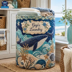May include: A cylindrical quilted laundry hamper featuring a dark blue whale, ocean waves, seashells, and starfish in shades of blue, teal, and tan. A central rectangular patch displays the embroidered text Gigi's Laundry surrounded by floral accents.