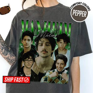 May include: Dark gray t-shirt featuring a graphic of five men with the text "MANHEIM Milo" in green. The shirt has a vintage look with a distressed design. The words "COMFORT COLORS" and "PEPPER" are printed on the upper right.