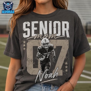 May include: A charcoal gray t-shirt featuring the word SENIOR in large white letters, the word mom in cursive, and a large number 07. A black and white graphic of a football player wearing a jersey with GRETNA 17 is centered on the shirt.