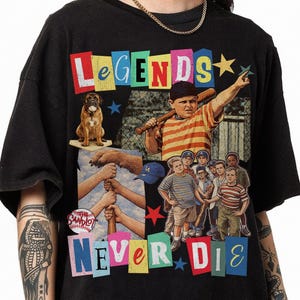 May include: Black t-shirt featuring a colorful graphic with the words "LEGENDS NEVER DIE." The design includes illustrations of a baseball team, a dog, and a baseball bat. The shirt has short sleeves and a crew neck.