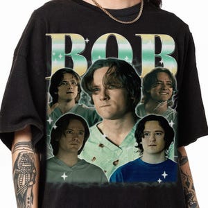 May include: Black t-shirt with a graphic print of four men's faces. The faces are surrounded by a yellow and green graphic that says "BOB".