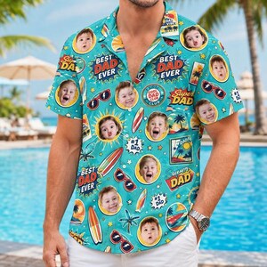 May include: A short-sleeve button-down shirt in teal featuring a repeating pattern of children's faces, surfboards, sunglasses, palm trees, and sunbursts. The text reads Best Dad Ever, Super Dad, and #1 Dad, with a collar and a front button closure.