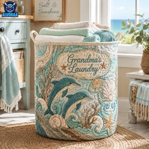 May include: A cylindrical fabric laundry hamper featuring a quilted design with two dolphins, seashells, and coral in shades of teal and beige. The front displays an embroidered label that reads Grandma's Laundry with two small starfish accents.