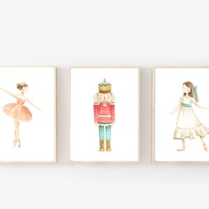 Puede incluir: Tres ilustraciones de acuarela enmarcadas. La primera ilustración presenta una bailarina con un tutú de color melocotón. La segunda ilustración presenta un cascanueces con un uniforme rojo y dorado. La tercera ilustración presenta una niña con un vestido blanco con una cinta azul.
